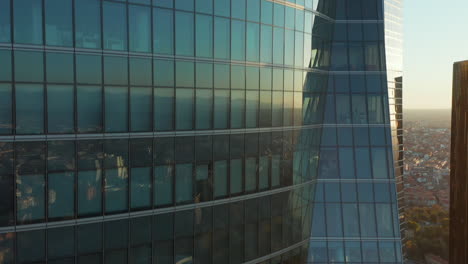 Ascending-footage-along-windows-and-glass-wall-of-office-skyscraper-reflecting-buildings-in-town-and-setting-sun.-Revealing-Spanish-flag-on-rooftop.
