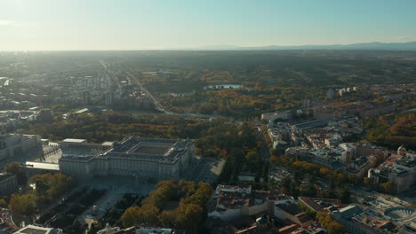 Panoramic-aerial-footage-of-historic-Royal-Palace-and-its-gardens-around.-Fly-above-city,-view-against-sun.