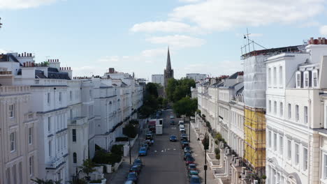 Forwards-fly-through-street-with-white-painted-houses.-Tall-church-spire-in-park-at-end-of-street.-London,-UK