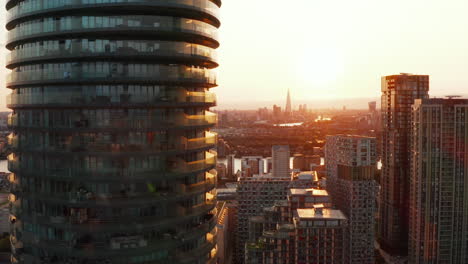 Fly-around-tall-modern-cylindrical-residential-building-Arena-Tower-in-Canary-Wharf-borough.-Futuristic-city-district-in-sunset-time.-London,-UK