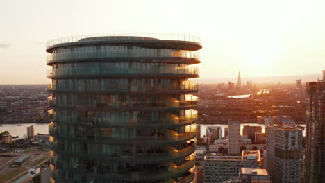 Pull-back-footage-of-futuristic-cylindrical-apartment-building-Arena-Tower-and-skyline-against-sunset-sky-in-background.-London,-UK