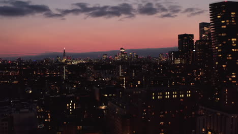 Landing-footage-of-city-lights-against-dusk-sky.-Evening-town-and-lighted-windows-of-various-buildings.-London,-UK