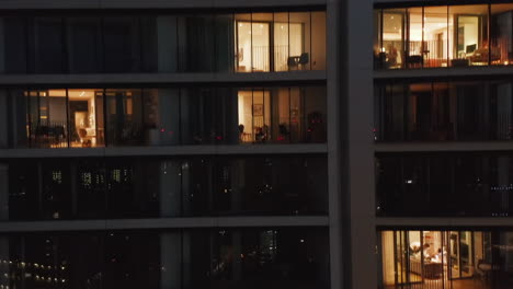 Crane-up-footage-of-high-rise-residential-building-at-night.-Close-up-view-of-wall-with-windows.-People-moving-inside-flats.-London,-UK