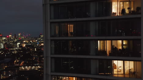 Descending-shot-along-wall-on-modern-high-rise-residential-building.-View-through-windows-into-luxury-flats.-Night-view-of-city.-London,-UK