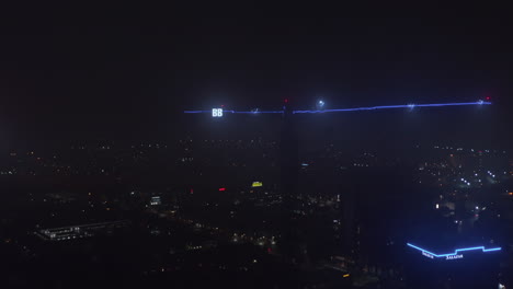 Arm-of-construction-crane-illuminated-with-blue-neon-light-and-two-big-letters-B.-Night-cityscape-with-lights-in-background.-Aerial-view-from-drone.-Dallas,-Texas,-US