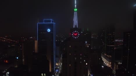 Colorful-Neon-illuminated-spike-and-clock-on-top-of-Mercantile-National-Bank-Building.-Aerial-drone-night-zooming-out-footage.-Dallas,-Texas,-US