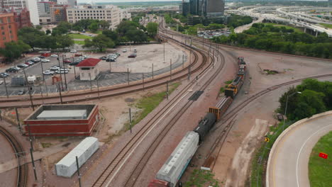 Aerial-view-of-freight-train-on-the-tracks.-Transport-of-good-on-railway-corridor-winding-between-highway-and-downtown.-Drone-following-rail-vehicles.-Dallas,-Texas,-US