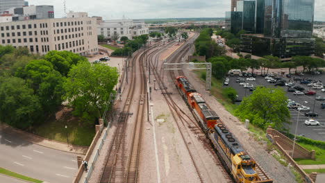 Three-powerful-diesel-engines-leading-freight-train-around-modern-glass-facade-skyscrapers.-Aerial-drone-view-of-slowly-running-train-arriving-to-station.-Dallas,-Texas,-US