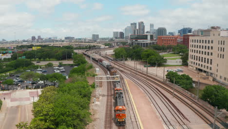 Aerial-view-of-cargo-train-slowly-riding-through-EBJ-Union-station.-Backwards-flying-drone-following-railway-corridor.-Skyline-with-skyscrapers-in-background.-Dallas,-Texas,-US