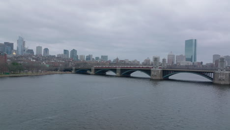 Red-line-train-driving-on-Longfellow-Bridge-with-urban-borough-in-background.-Forwards-fly-above-rippled-water-surface.-Boston,-USA