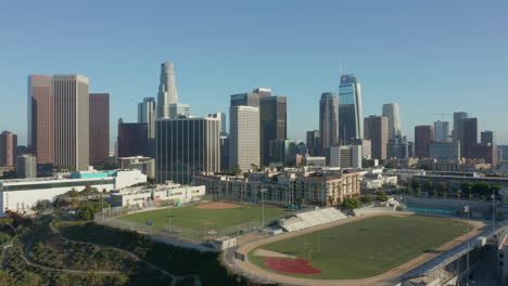 Empty-Sports-Stadium-by-City-Skyline,-Aerial-Dolly-in-revealing-Skyscrapers-of-Los-Angeles,-California,-United-States