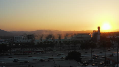 AERIAL:-Steaming-Factory-with-Palm-Trees-and-busy-Highway-in-Burbank,-Los-Angeles,-California,-Sunset