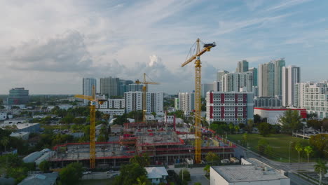 Slide-and-pan-footage-of-cranes-on-construction-site-at-twilight,-urban-development-in-background.-Miami,-USA