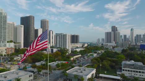 Aerial-view-of-urban-borough-with-buildings-and-greenery.-Backwards-reveal-of-US-flag-and-high-rise-apartment-buildings-in-background.-Miami,-USA
