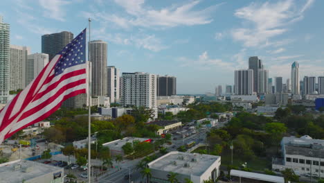 Fly-around--American-national-flag-waving-on-pole-above-city.-High-rise-office-or-apartment-buildings-in-background.-Miami,-USA