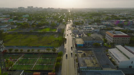 Backwards-fly-above-long-straight-and-wide-street-leading-in-urban-neighbourhood.-Golden-hour-in-city.-Miami,-USA