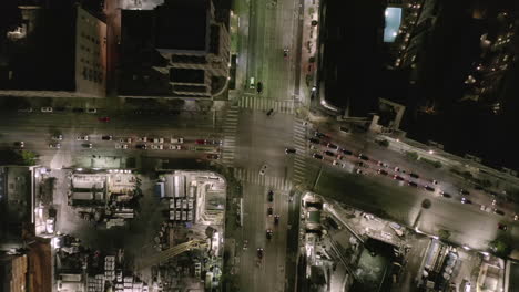 AERIAL:-Overhead-Rising-on-Intersection-Street-with-big-Construction-Site-and-Holes-in-Ground-at-Night-with-Glowing-Streets-and-City-Car-Traffic-Lights