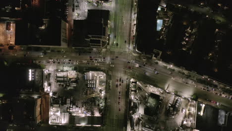 AERIAL:-Overhead-Rising-on-Intersection-Street-with-big-Construction-Site-and-Holes-in-Ground-at-Night-with-Glowing-Streets-and-City-Car-Traffic-Lights