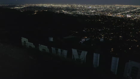 Antenne:-Spektakulärer-Flug-Nah-über-Hollywood-Sign-Letter-Bei-Nacht-Mit-Lichtern-Im-Stadtbild-Von-Los-Angeles