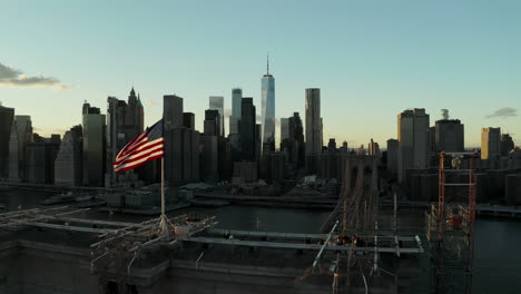 Aerial-view-of-historic-bridge-spanning-river-and-cityscape-with-modern-skyscrapers.-Backwards-reveal-of-stone-suspension-tower-with-American-flag-on-top.-Manhattan,-New-York-City,-USA