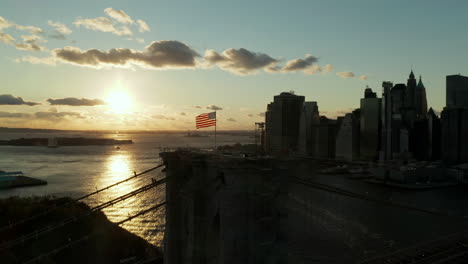 Orbit-shot-around-American-flag-waving-in-wind-on-top-of-Brooklyn-Bridge.-Silhouette-of-downtown-skyscraper-in-Financial-District-against-sunset-sky.-Manhattan,-New-York-City,-USA