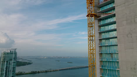 Fly-around-construction-of-new-skyscraper.-Revealing-panoramic-view-of-sea-bay-and-aircraft-in-sky.-Miami,-USA