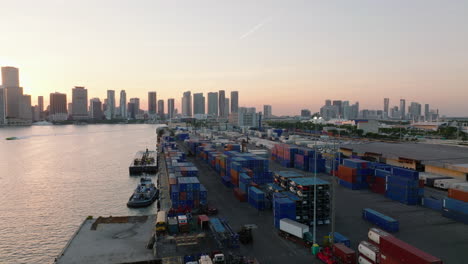 Fly-above-stacks-of-naval-containers-in-harbour-on-Dodge-Island.-Silhouette-on-high-rise-downtown-buildings-against-sunset.-Miami,-USA
