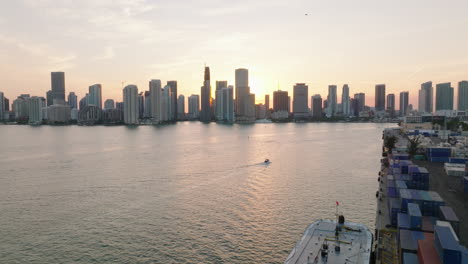 Panoramic-view-of-modern-downtown-buildings-on-waterfront-against-sunset-sky.-Backwards-reveal-naval-containers-and-ship-in-harbour.-Miami,-USA
