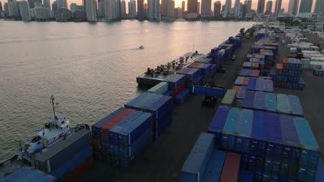 Forwards-fly-above-rows-of-stacked-overseas-containers-in-harbour.-Tilt-up-reveal-skyline-against-setting-sun.-Miami,-USA