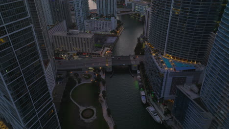 Backwards-fly-above-river-in-city.-High-angle-view-of-road-bridge-and-yachts-moored-at-banks,-high-rise-buildings-on-waterfronts.-Miami,-USA