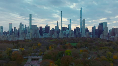 Wide-panoramic-view-of-skyscrapers-surrounding-Central-park.-Fly-above-autumn-colour-trees-in-park-at-dusk.-Manhattan,-New-York-City,-USA
