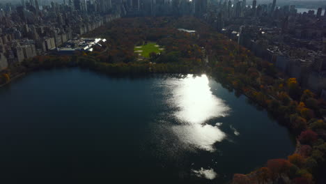 Aerial-view-of-shimmering-water-surface-in-autumn-Central-Park.-Tilt-up-reveal-of-modern-business-skyscrapers.-Manhattan,-New-York-City,-USA