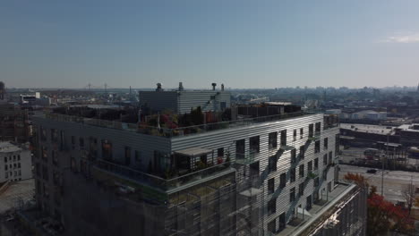 Fly-over-apartment-building-with-rooftop-terraces-and-scaffolding-around-facade.-Revealing-multilane-expressway-and-panoramic-view-of-industrial-or-logistic-site.-New-York-City,-USA
