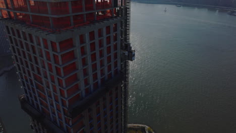 Aerial-descending-footage-of-high-rise-building-construction-site-on-riverbank.-Downtown-skyscrapers-in-distance.-New-York-City,-USA